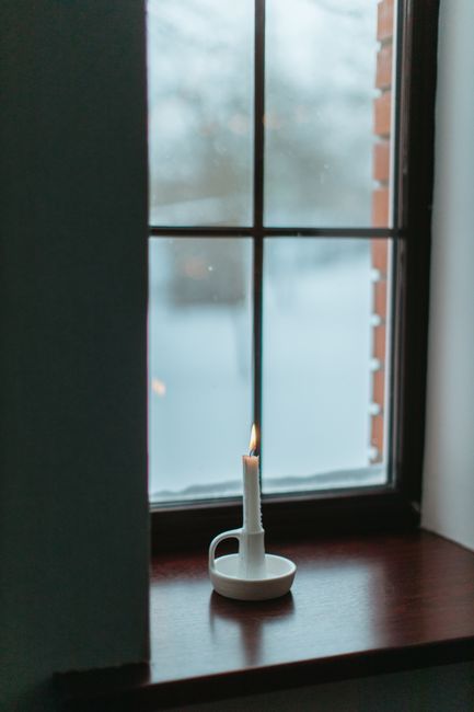 A lit candle in a holder sits on a window sill, casting a warm glow by the window with a snowy view.