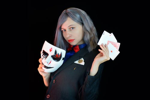 Side view of serious young female casino dealer in classy suit with bow tie holding face mask and cards in hands and looking at camera against black background