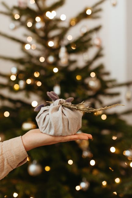 A hand holding a wrapped Christmas gift in front of a blurred decorated tree.