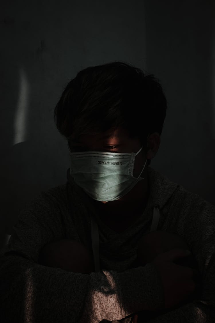 Sad Asian Man In Mask Looking Away While Sitting On Floor And Embracing Knees