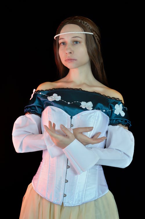 Beautiful young female model wearing elegant medieval corset dress and pearl diadem crossing hands gracefully and looking away against black wall in photo studio