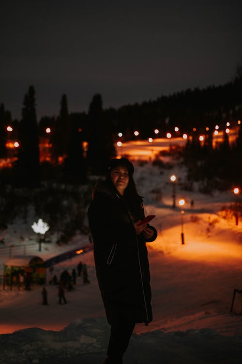 Woman in Black Coat Standing on Snow Covered Ground during Night Time