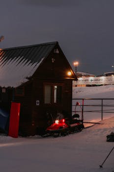 Snow-covered cabin illuminated at night with a snowmobile outside, creating a winter scene.