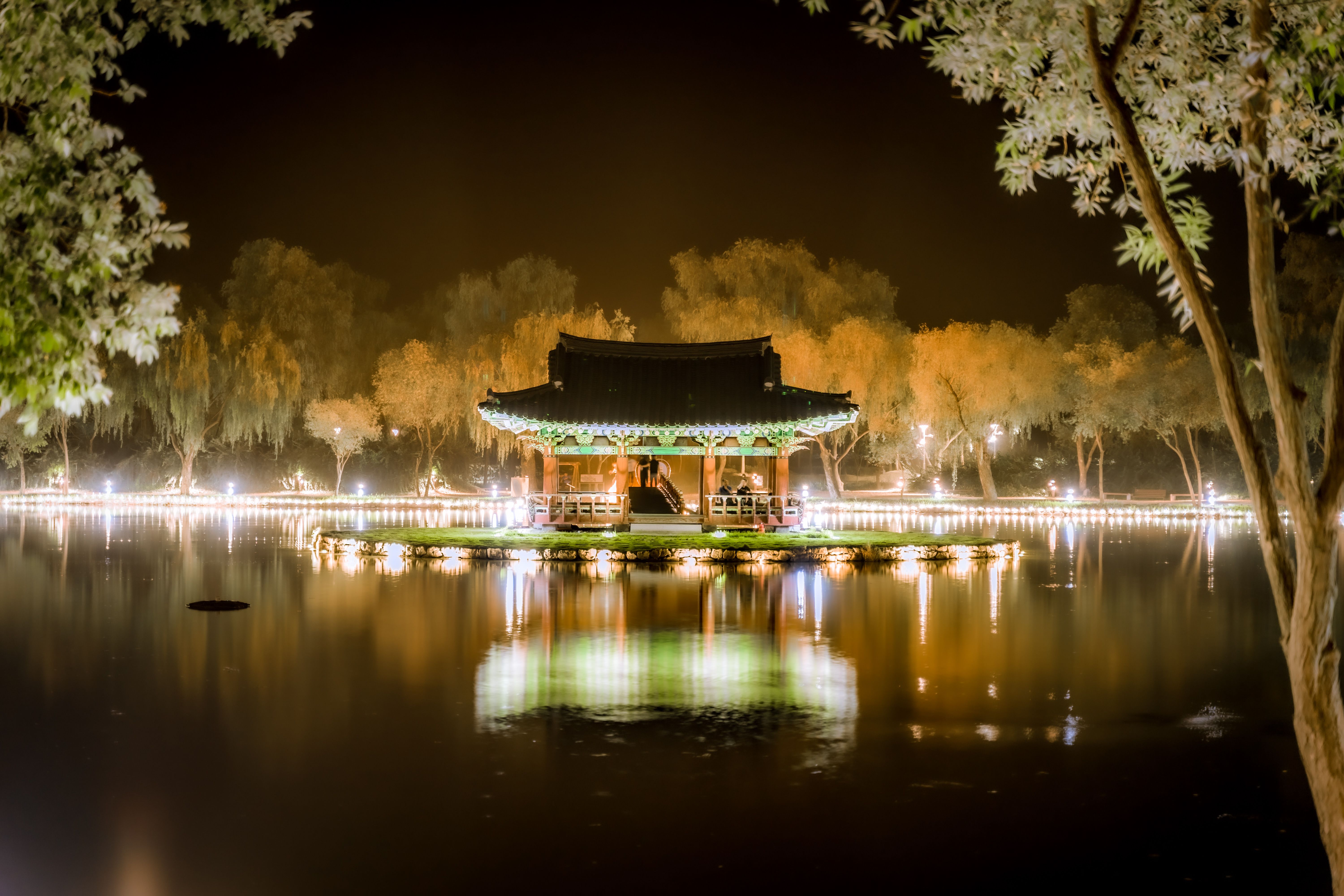 A serene night view of an illuminated gazebo surrounded by water and trees in a South Korean park.
