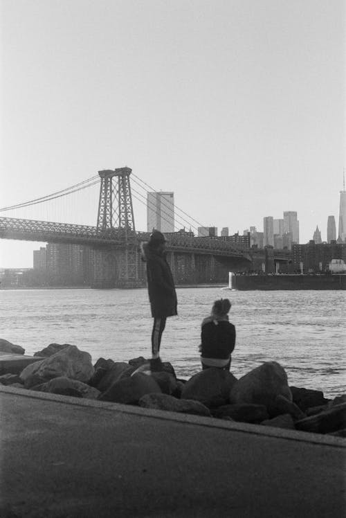 Graustufenfoto Von Mann Und Frau, Die Auf Rocky Shore Nahe Brücke Gehen