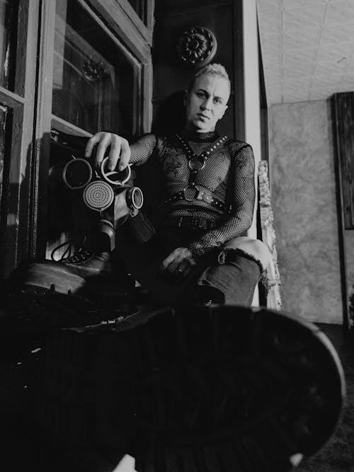 Black and white of male in harness sitting at home near gas mask and looking at camera
