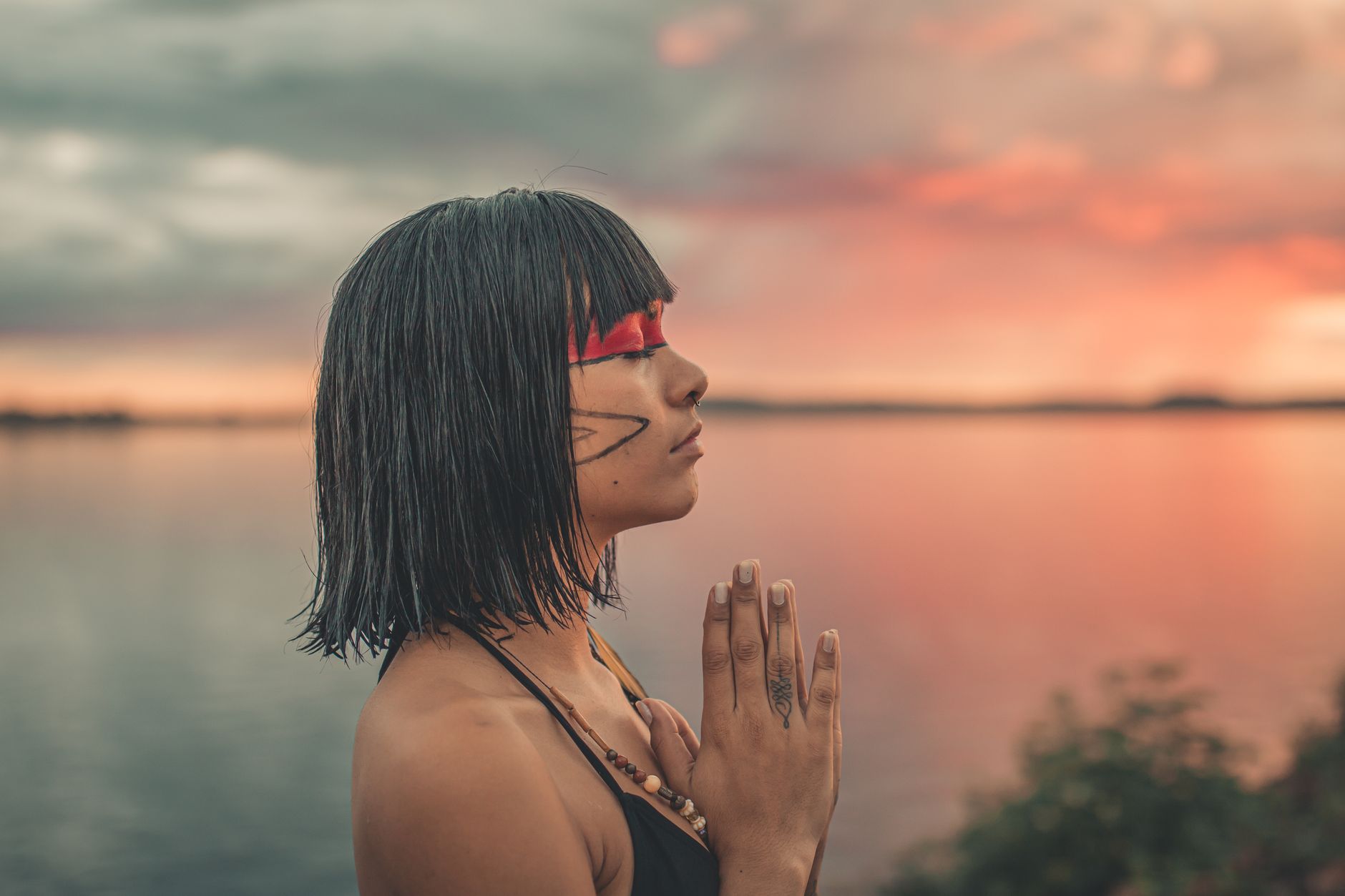 Woman meditating peacefully by the lake at sunset with face paint and closed eyes.