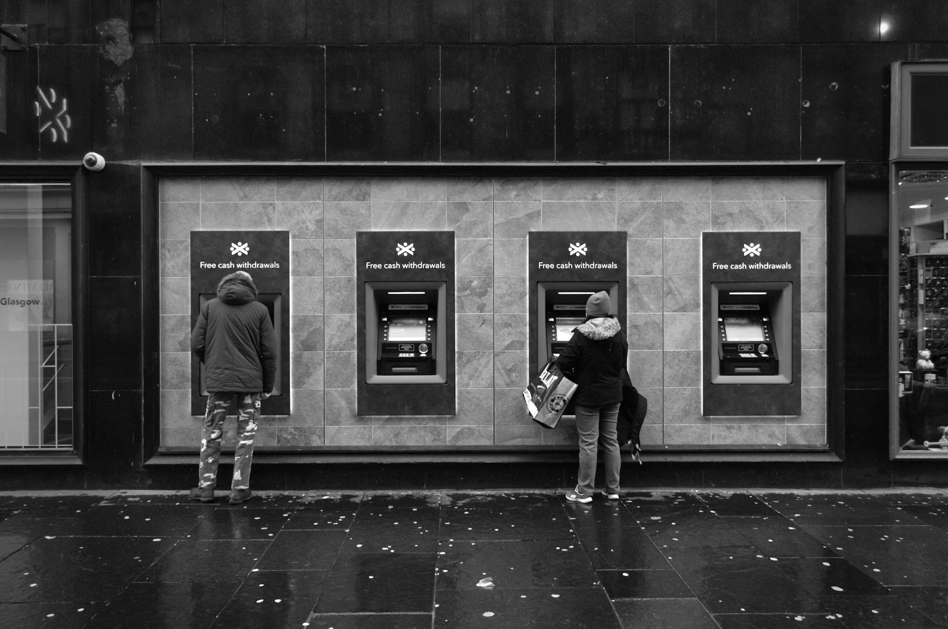 Black and white photo of people using ATMs in an urban setting, showing modern city life.