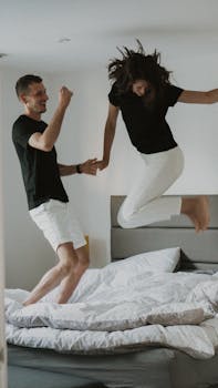 A joyful couple jumping on a bed in a well-lit bedroom, holding hands and having fun.
