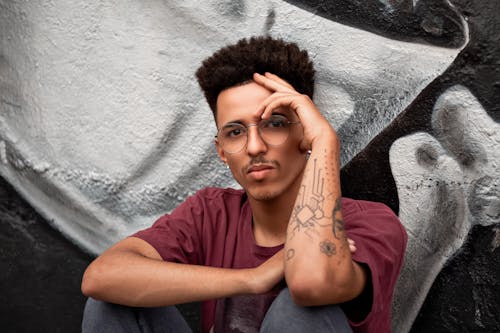 Serious young Hispanic male in eyeglasses with tattoo sitting near wall with graffiti and looking at camera touching face