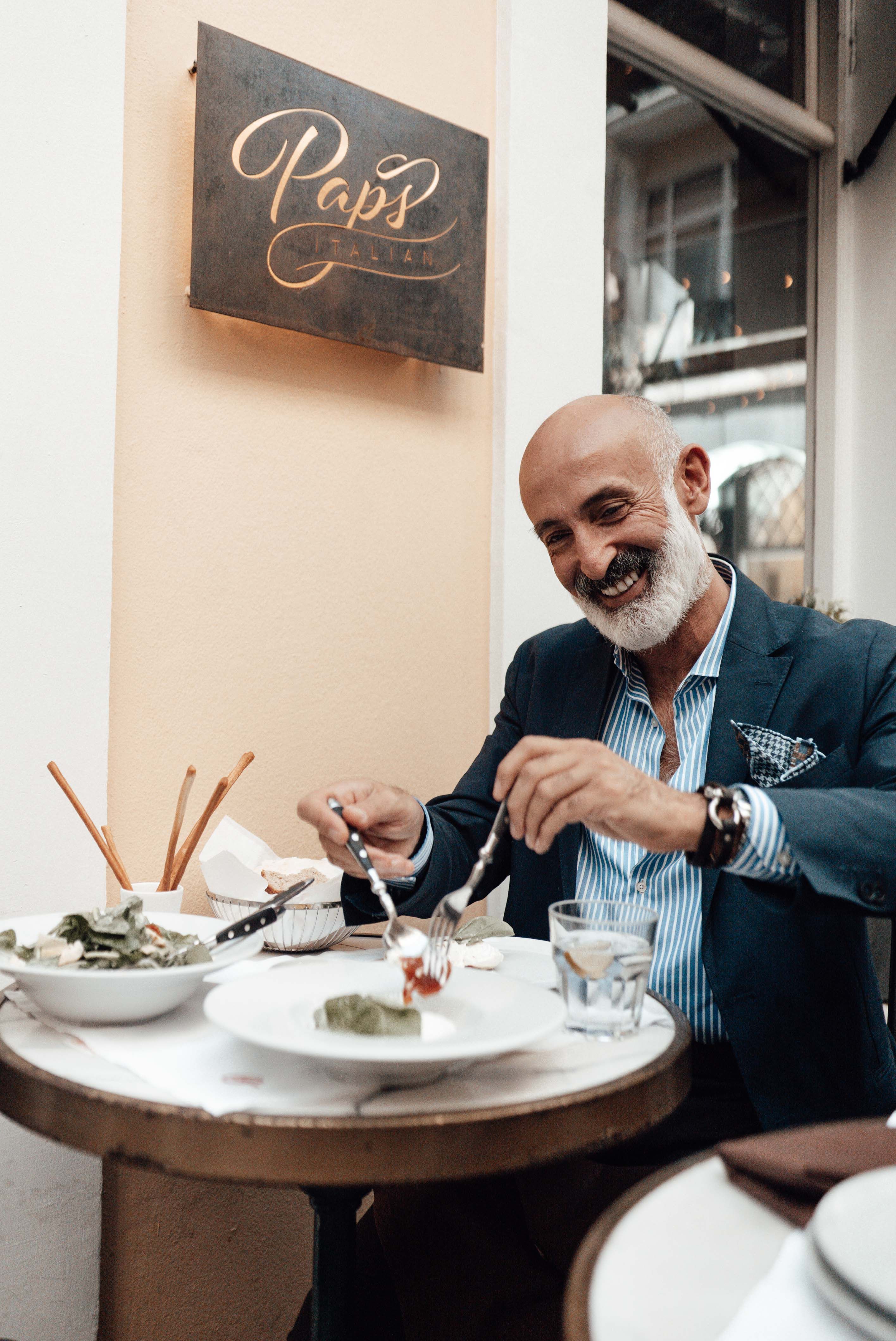 Cheerful stylish bearded ethnic elderly male in elegant suit smiling while sitting at table on cafe terrace and eating delicious dishes during lunch