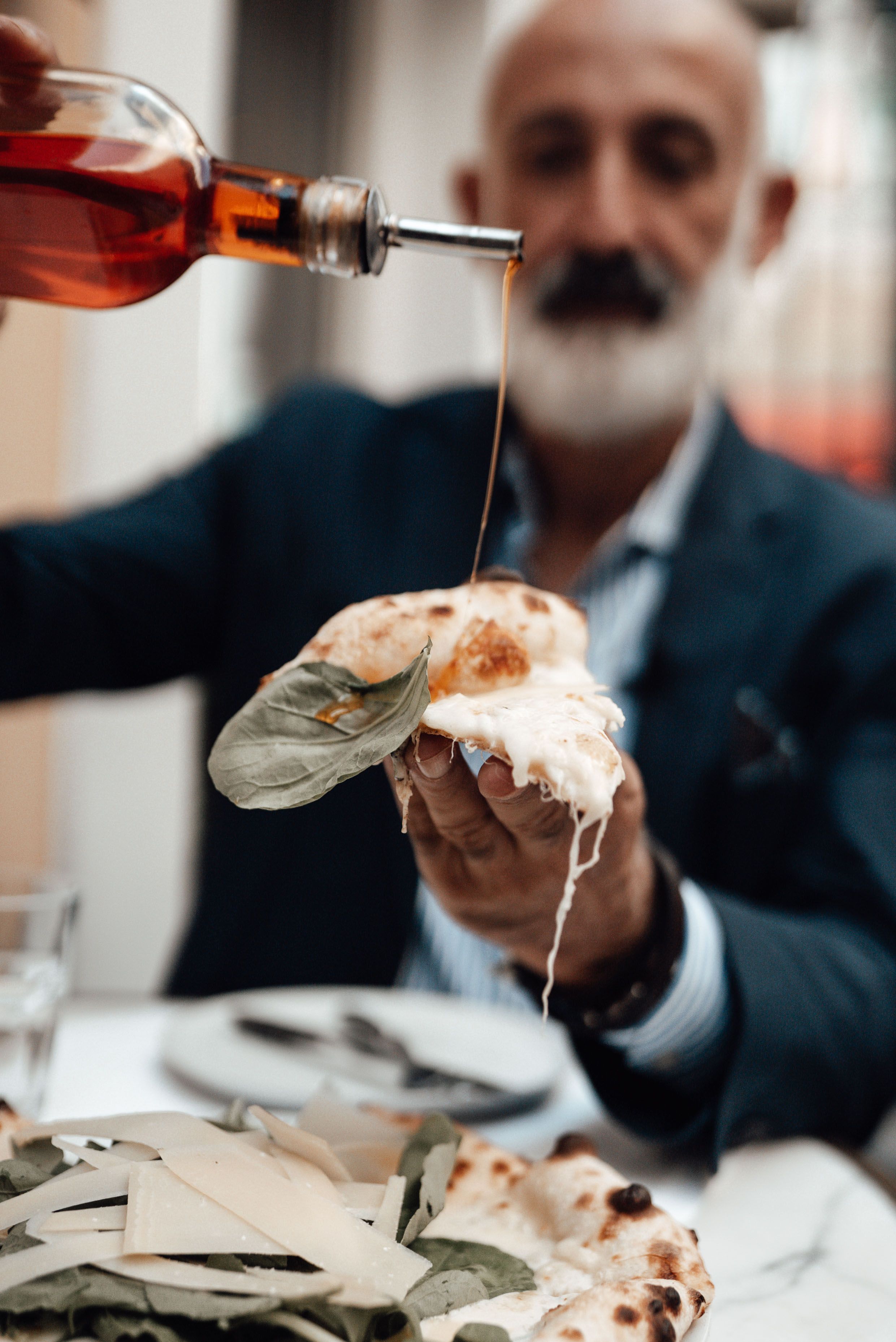 Stylish senior bearded ethnic male in suit pouring vinegar on slice of pizza while having lunch in restaurant