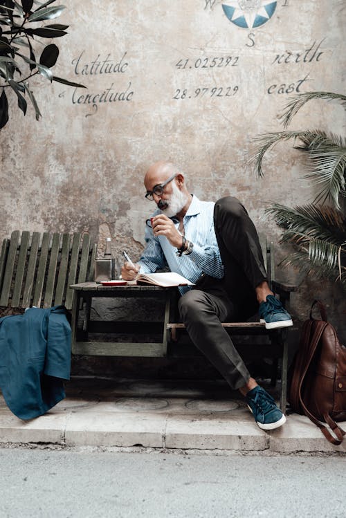 Full body of concentrated senior bearded ethnic male author in stylish clothes and eyeglasses drinking coffee and writing new article in notebook while relaxing in outdoor cafe