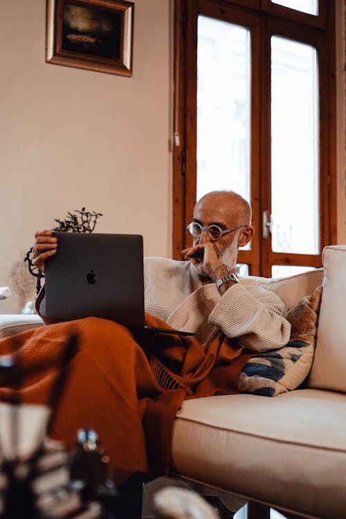 Concentrated elderly bearded ethnic male freelancer in robe and eyeglasses sitting on comfortable sofa and working remotely on laptop at home