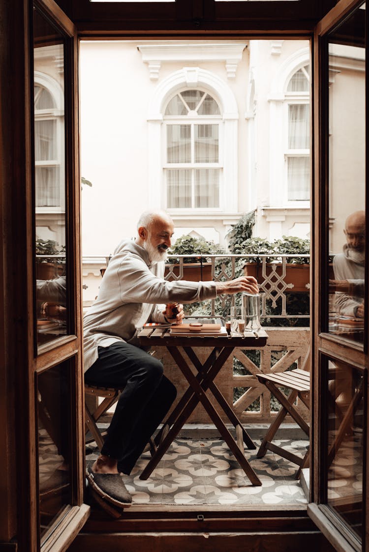 Smiling Elderly Ethnic Man Resting At Table On Balcony At Home