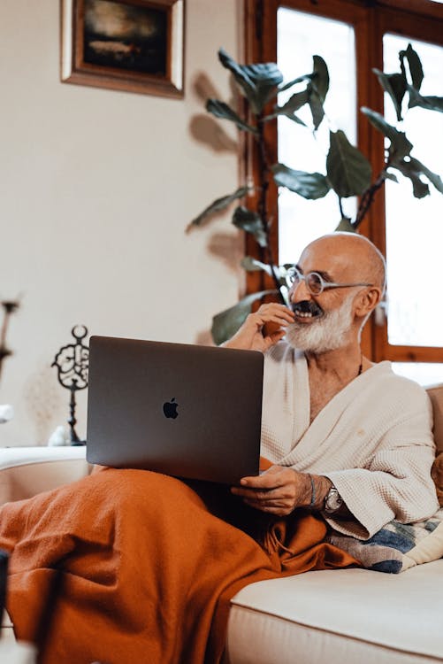 Joyful elderly ethnic male with white beard in bathrobe smiling while relaxing on comfortable sofa and watching funny movie on laptop during weekend at home