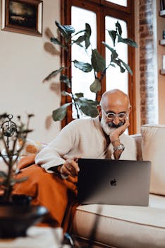 Elderly man in bathrobe using laptop at home, enjoying a cozy lifestyle in living room.