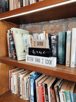 Collection of assorted books and decorative wooden elements with inscription I drink vine better than i cook placed on shelves in bookstore in daytime