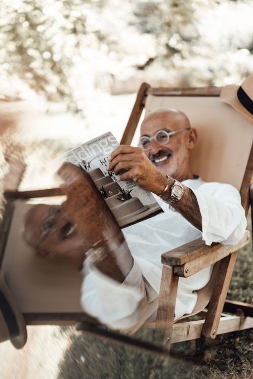 Cheerful senior ethnic man in eyeglasses and stylish clothes sitting on chair and reading magazine while resting in nature among green trees in sunny day