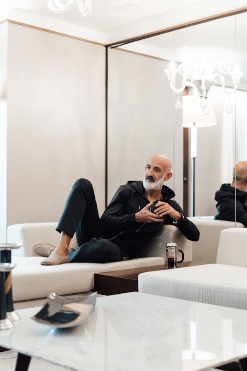 Full body of pensive senior bearded ethnic male in casual clothes relaxing on comfortable couch near mirrored wall and drinking hot tea in modern apartment