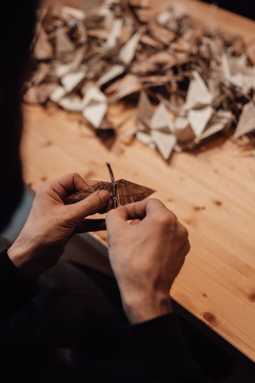 High angle of crop anonymous male craftsman creating origami crane near wooden table with many various figurines from paper in light workshop