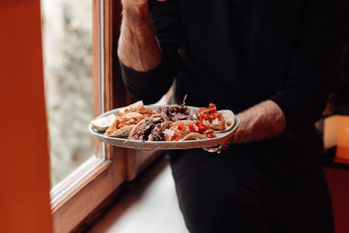 Crop faceless male in black casual clothes sitting on windowsill near window with plate with tasty tacos with onions and tomatoes with chicken near lime in daylight in room