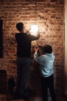 Back view of anonymous guys in casual clothes standing in cafe near brick wall while hanging big garland with dry herbs and lamp