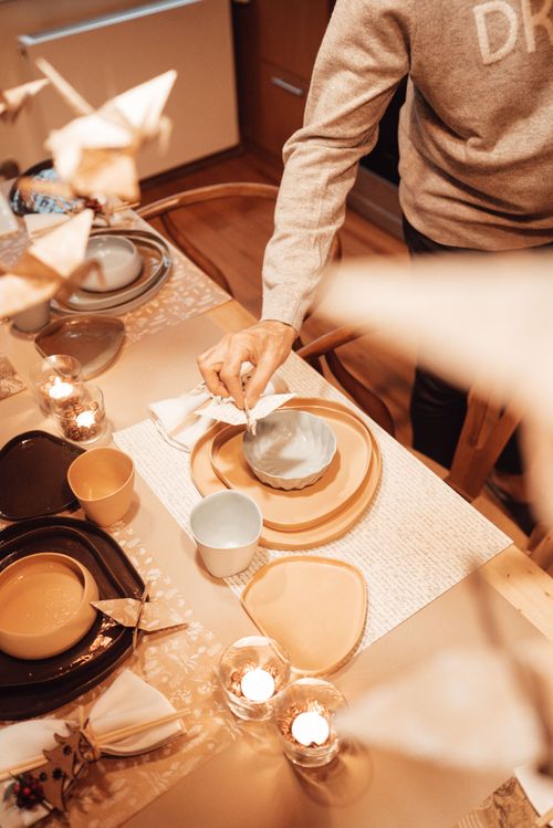 Free From above of unrecognizable male putting paper crane on served table with various dinnerware and burning candles in light kitchen Stock Photo