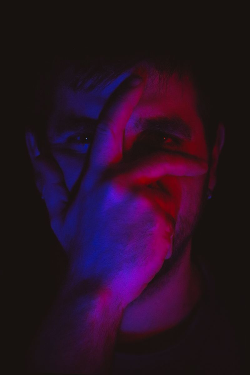 A close-up of a man with his hand covering his face, illuminated by moody lighting.