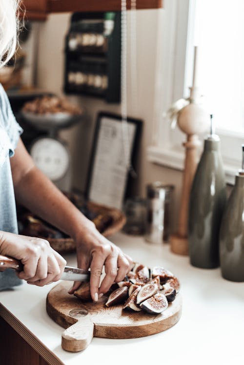 Unrecognizable female cook with knife cutting heap of figs on wooden cutting board while standing at kitchen counter on blurred background