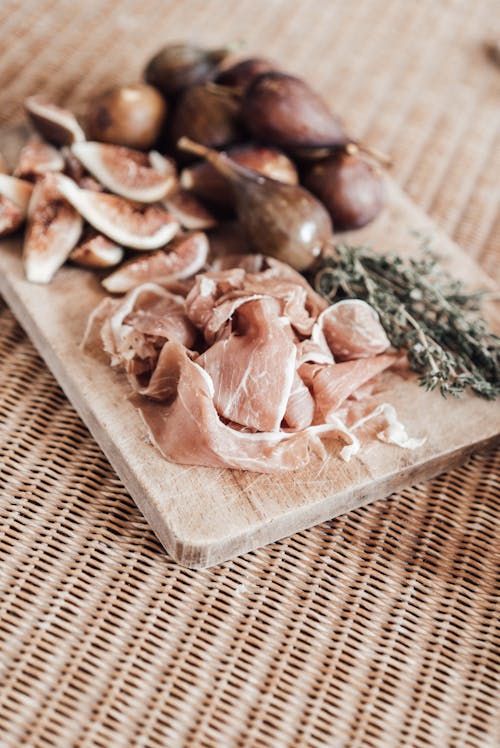 Free Slices of fresh ham on wooden board with whole and cut ripe figs near dried thyme on table in kitchen Stock Photo