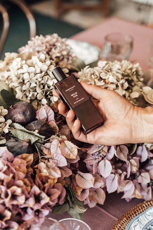 High angle of crop anonymous person with bottle of perfume under delicate fragrant flowers placed on table for occasion