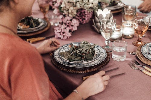 High angle side view of crop female with portion of salad sitting at festive table decorated with blooming flower and burning candles