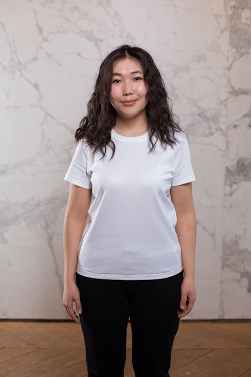 Glad young Asian female wearing black pants and white t shirt standing calmly against marble wall in light room and looking at camera