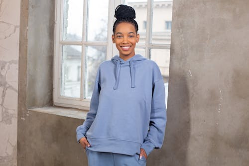 Cheerful attractive African American female wearing blue cozy hoodie standing with hands in pockets and looking at camera while standing near window in light room