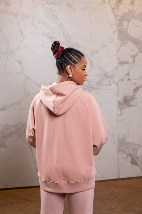 Back view of young African American female millennial with traditional braids in stylish pink hoodie standing in light room and looking away