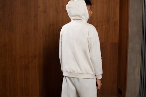 Back view of young ethnic stylish male model with Afro hair in trendy hooded sweatshirt standing near wooden wardrobe and looking away thoughtfully