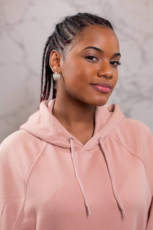 Self assured young black female model with Afro braids and perfect skin in stylish pink hoodie smiling while looking at camera against gray background