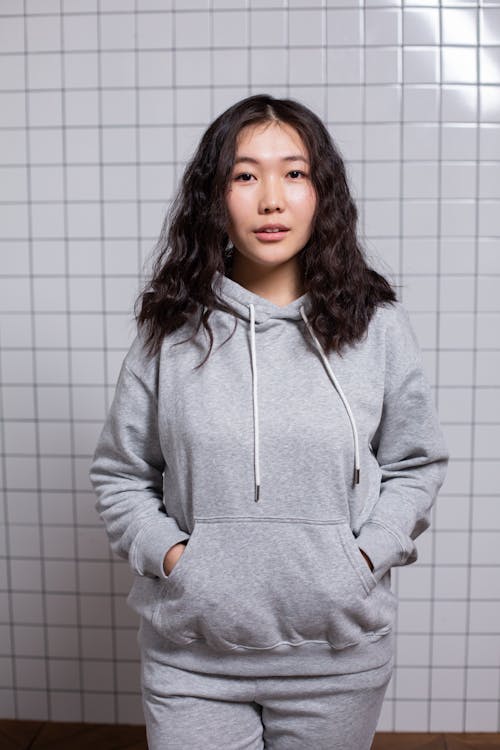 Positive Asian female teenager with dark wavy hair standing with hands in hoodie pocket and looking at camera in light room with tiled wall