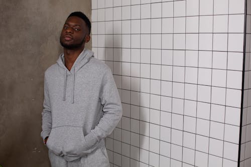 Confident African American male wearing gray hoodie and pants looking at camera while standing with hands in pockets near tiled wall