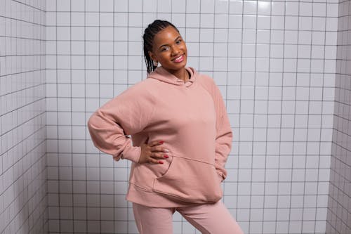 Positive young African American female in fashionable casual hoodie smiling and looking at camera near tiled wall