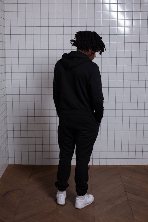 Back view of anonymous African American male in trendy black hoodie and trousers near tiled wall