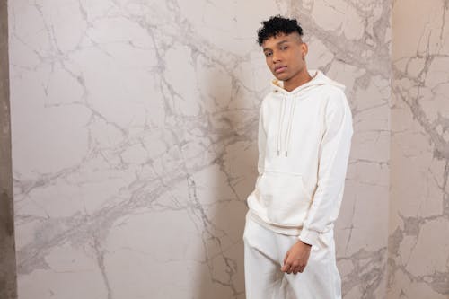 Confident black man with curly hair wearing white sports suit standing against marble wall and looking at camera