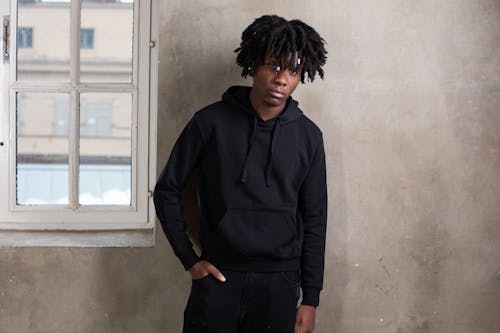 Confident African American man with dreads in casual black outfit standing in front of bare wall with small window and looking away