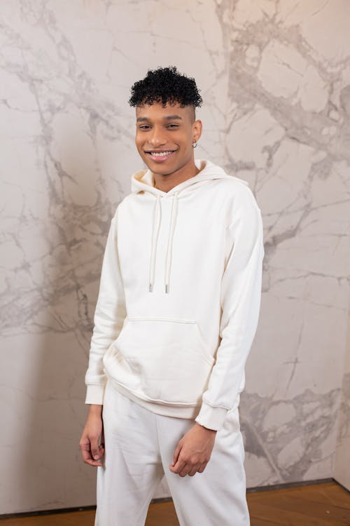 Positive African American male wearing trendy white hoodie and pants standing near wall with marble ornament and friendly looking at camera