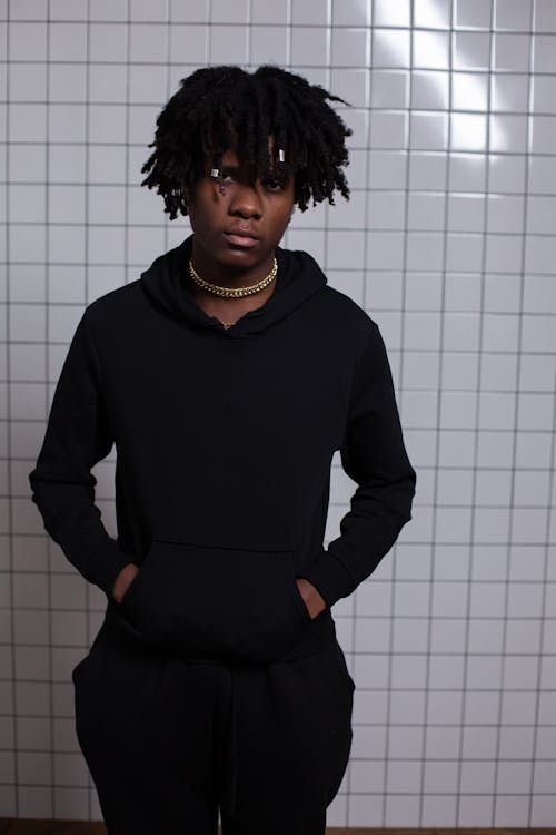 Confident young black male with dreadlocks putting hands in pockets of casual black hoodie and standing in front of tiled white wall