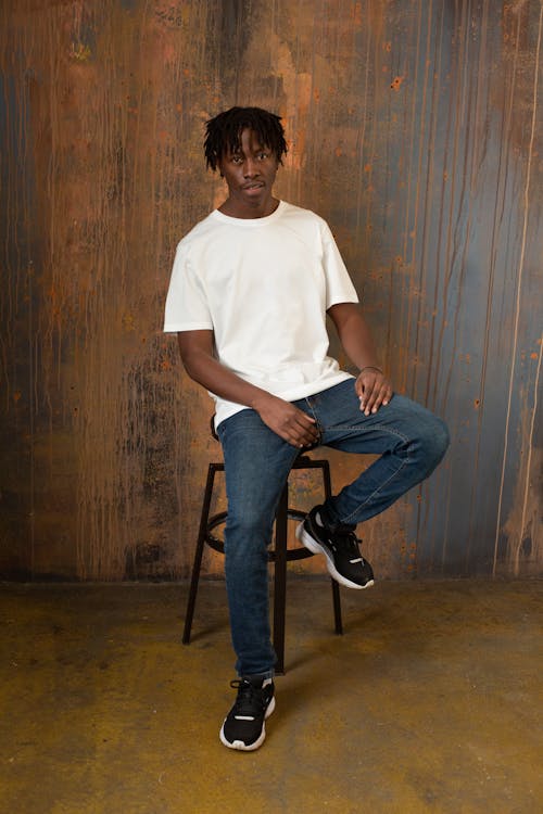 Full body of self assured young African American male model with dreadlocks in casual white t shirt and jeans sitting on chair and looking at camera against wooden wall