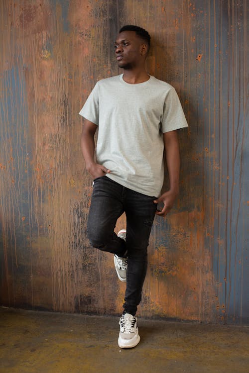 Full length of thoughtful young bearded African American guy in casual outfit leaning leg on wooden wall while standing in room and looking away