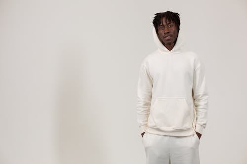Confident of young African American male millennial with dreadlocks in stylish hoodie standing against white background with hands in pockets and looking at camera