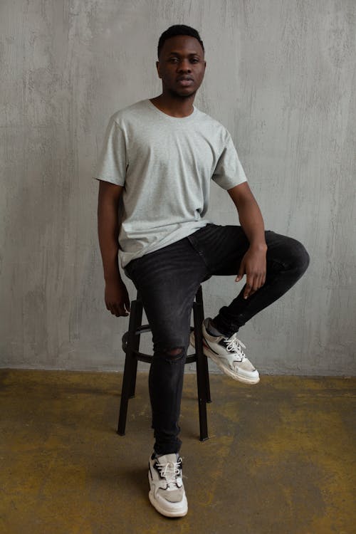 Full body of fashionable young African American guy in stylish casual outfit sitting on stool and looking at camera in studio